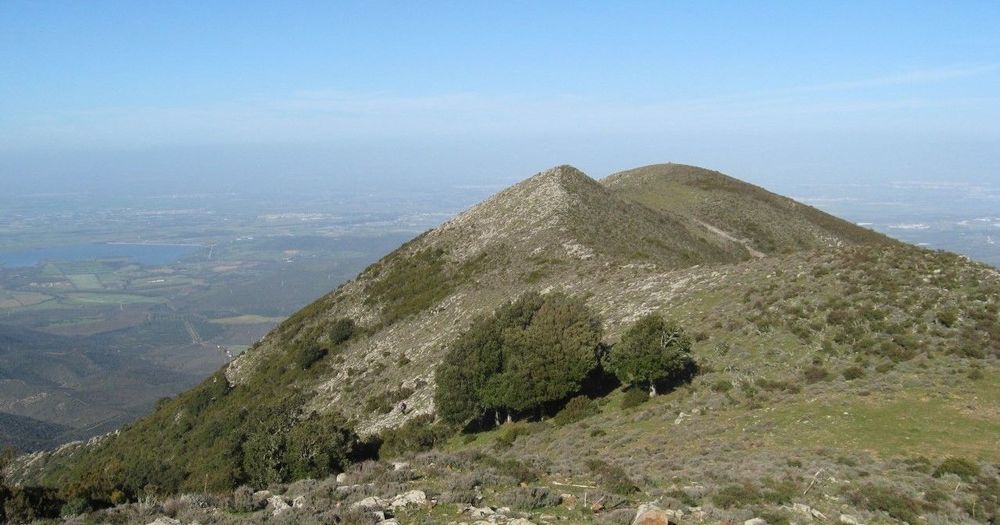 Veduta aerea di una montagna rocciosa e erbosa con alberi sparsi, sullo sfondo un lago e una valle verde sotto un cielo azzurro
