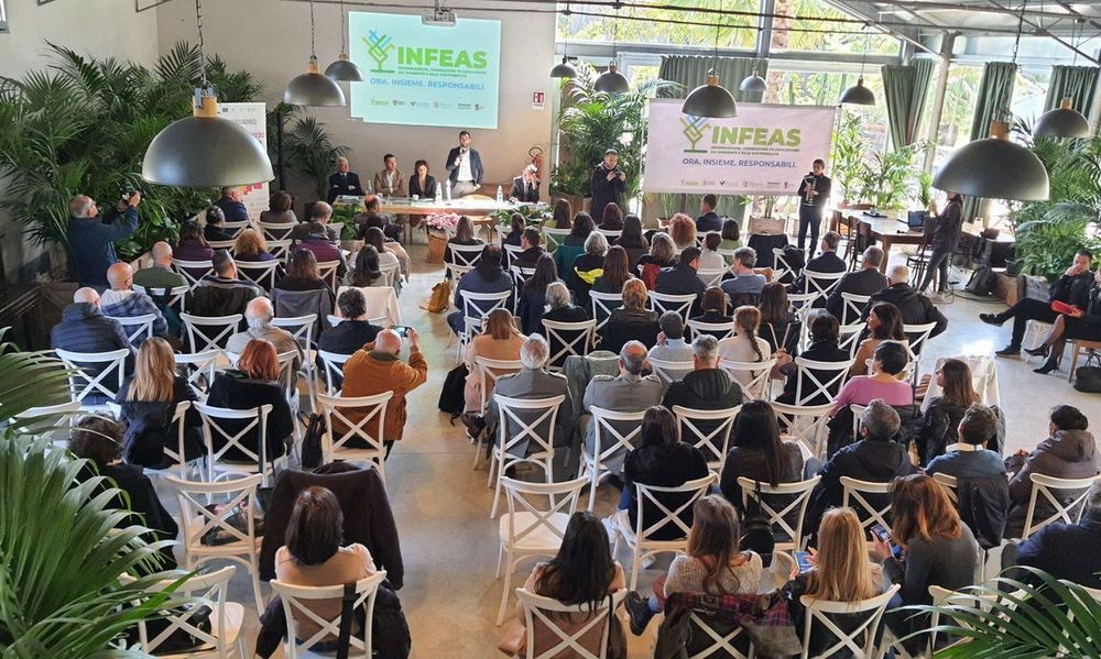 Una sala piena di persone sedute su sedie bianche, con un palco e uno schermo che mostra il logo INFEAS durante un evento.