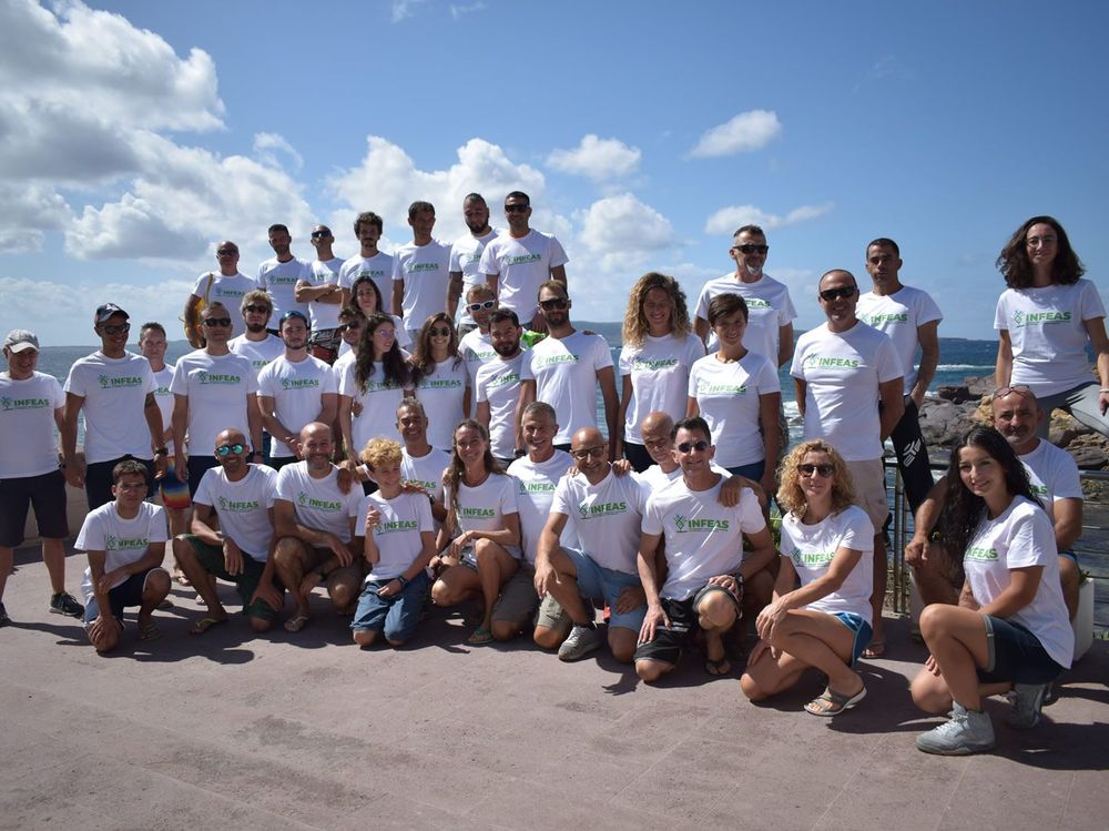 Gruppo di persone con maglietta bianca con logo INFEAS, in posa su un lungomare roccioso con vista sul mare e cielo azzurro con nuvole.