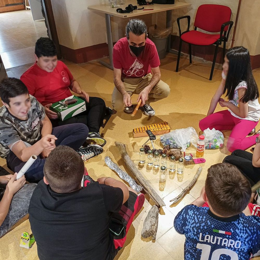 Un gruppo di bambini e un adulto siedono in cerchio per terra, circondati da vari oggetti come bottiglie, legni e altri materiali, probabilmente per u