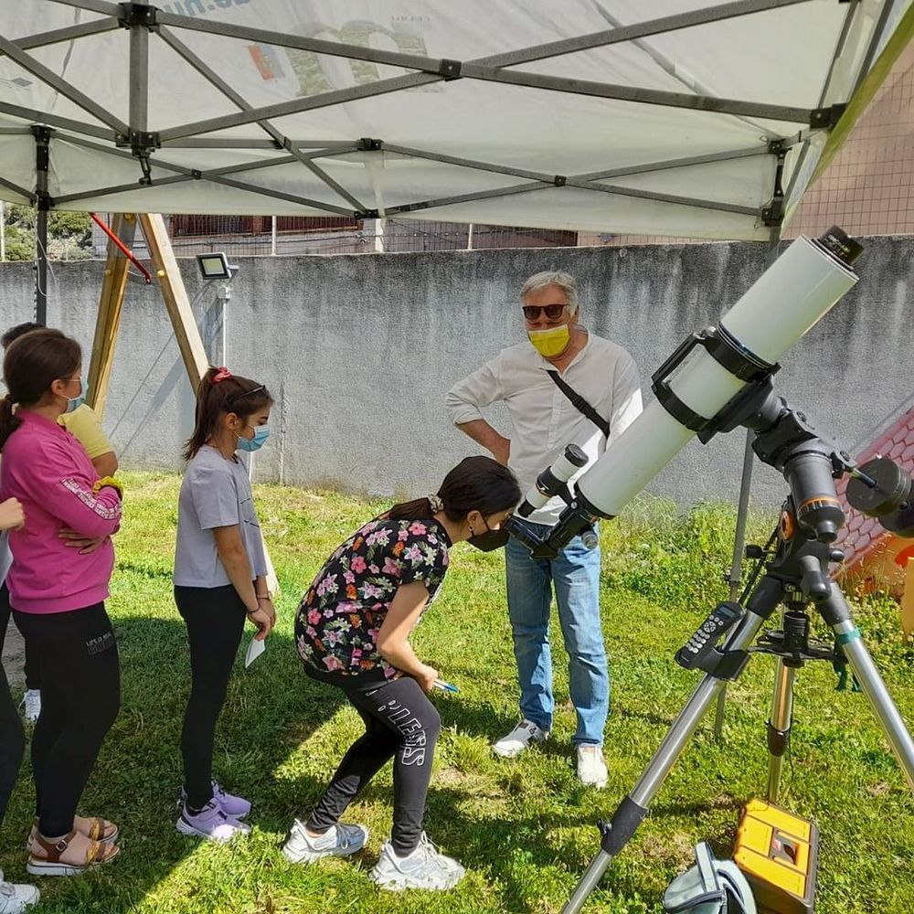 Un gruppo di persone, tra cui bambini e adulti, si riunisce intorno a un telescopio posizionato su un treppiede sotto un gazebo, tutti indossano masch