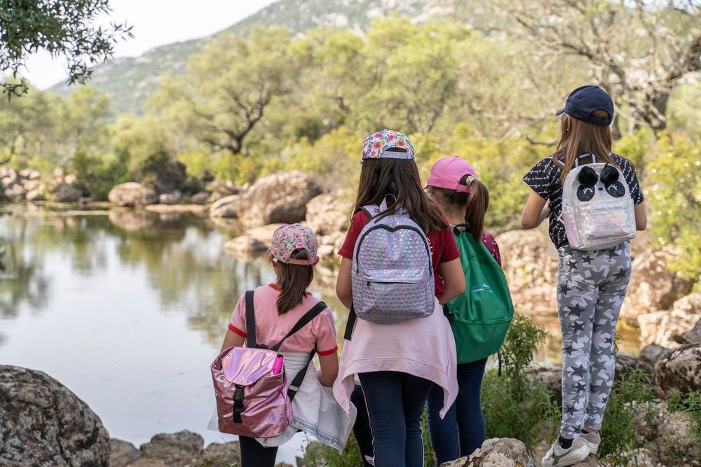 Quattro ragazze con zaini colorati guardano un lago circondato da alberi e rocce, con una montagna sullo sfondo.