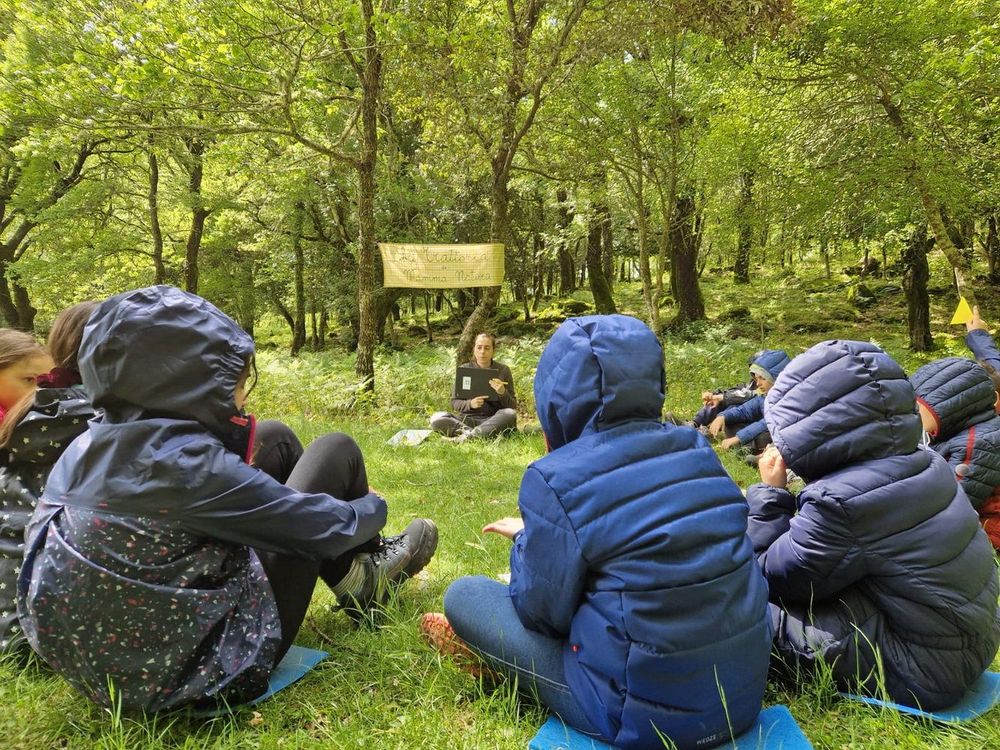 Gruppo di bambini seduti in cerchio in un bosco, ascoltano una donna che parla con un laptop, indossano giacche e sono su stuoie colorate.