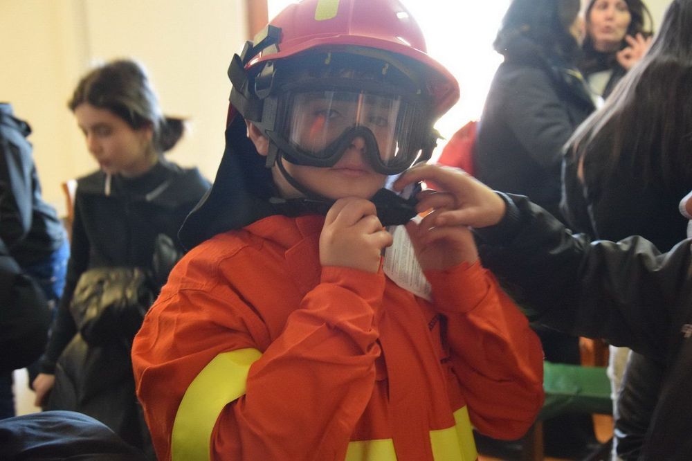 Un bambino indossa una tuta da pompiere arancione con strisce gialle e un casco rosso mentre una persona gli aggiusta la maschera nera sul viso.