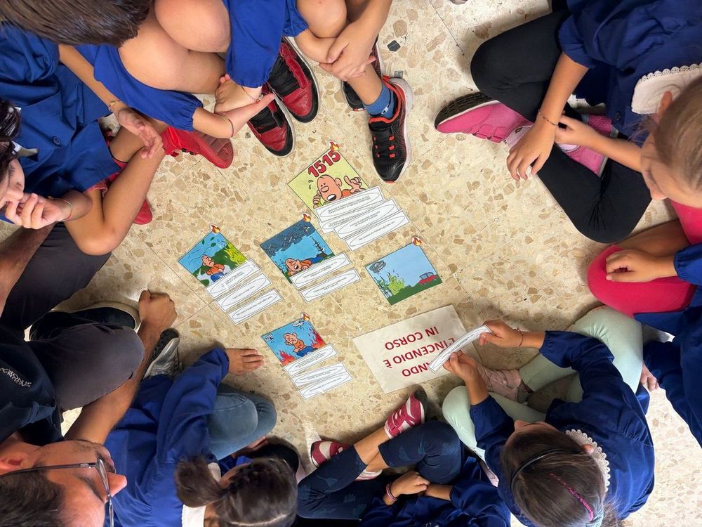 Bambini seduti in cerchio con carte colorate e un cartellone che dice 'Incendio in corso'