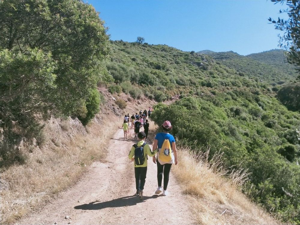 Un gruppo di persone, tra cui un bambino e una donna, camminano su un sentiero sterrato in una zona collinare con vegetazione.