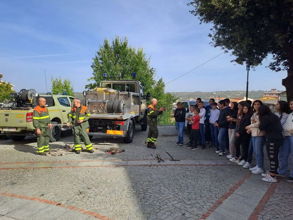 Vigili del fuoco in uniforme verde e arancione mostrano attrezzature a un gruppo di giovani in piedi su un selciato, con veicoli e alberi sullo sfondo