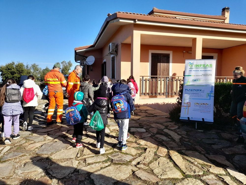 Gruppo di bambini e adulti, tra cui due uomini in divisa arancione, davanti a una casa con un cartellone informativo.
