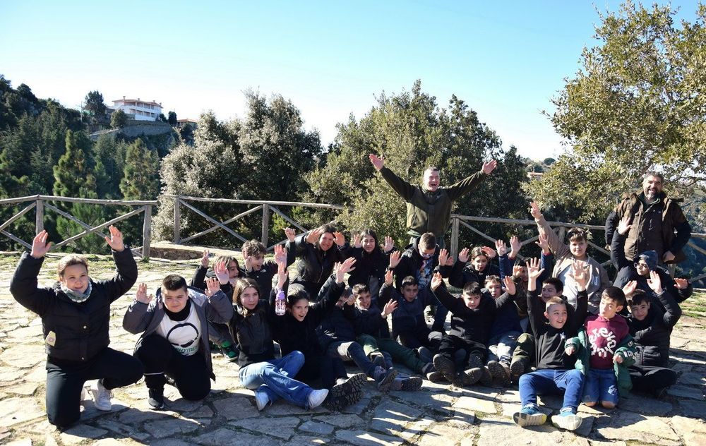 Gruppo di persone, prevalentemente bambini, che si riuniscono all'aperto, su un sentiero in pietra, con le mani alzate, circondati da alberi e un pano
