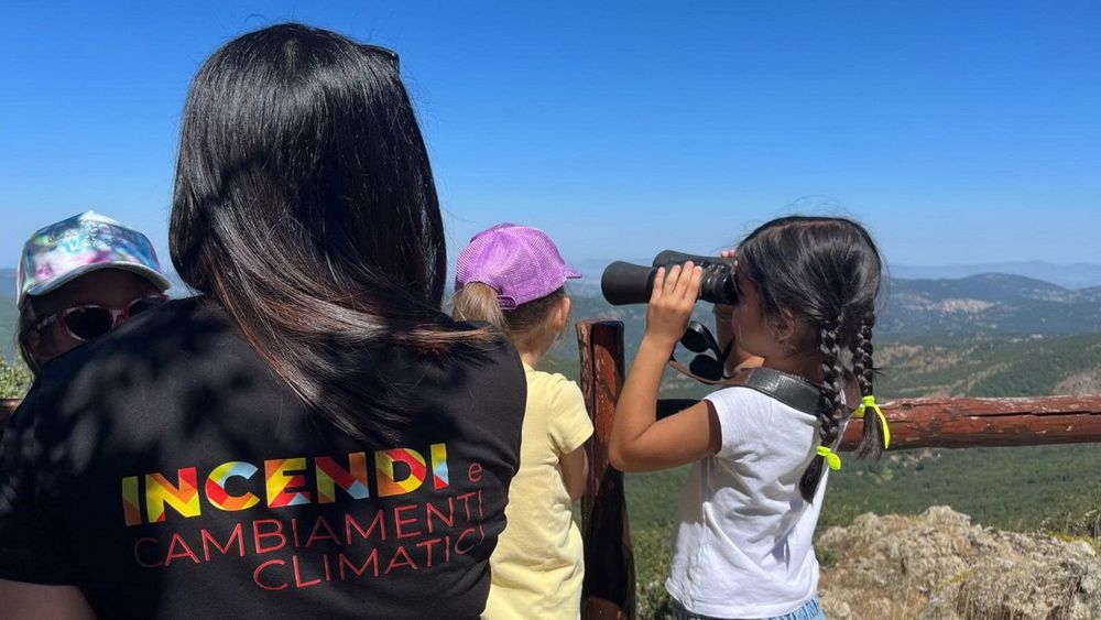 Tre persone, due adulti e una bambina, guardano un paesaggio naturale con un binocolo, con una maglietta che recita 'Incendi e cambiamenti climatici'.