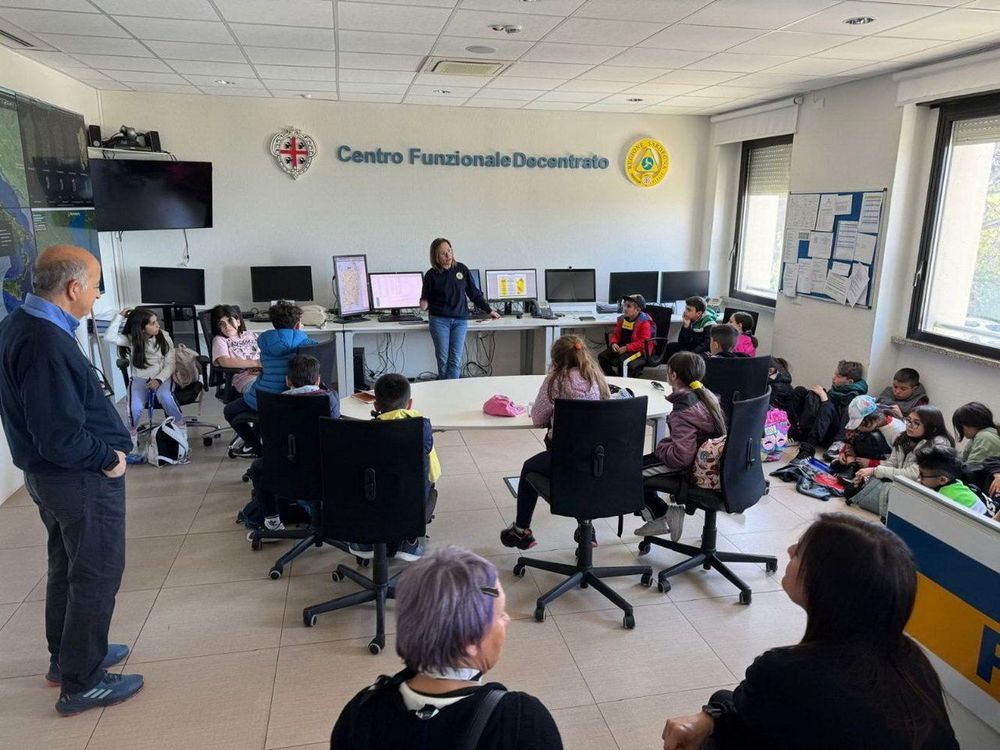 un'aula con bambini seduti intorno a un tavolo di fronte a un'insegnante che parla, con adulti in piedi, monitor e cartelli murali