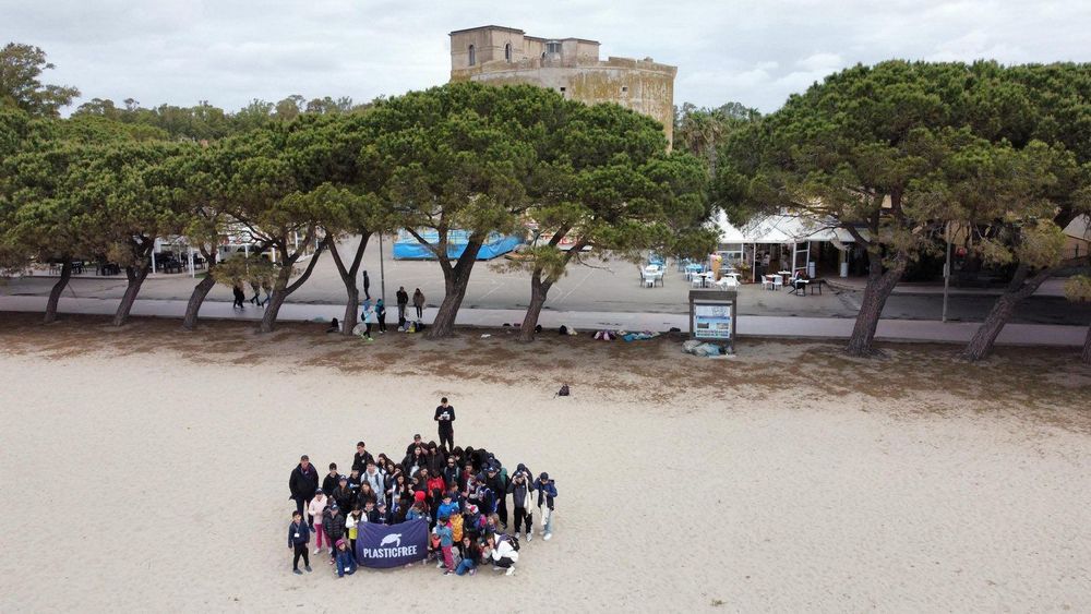 Un gruppo di persone di varie età, vestite con abiti caldi, si riunisce sulla spiaggia tenendo uno striscione con la scritta Plastic Free.