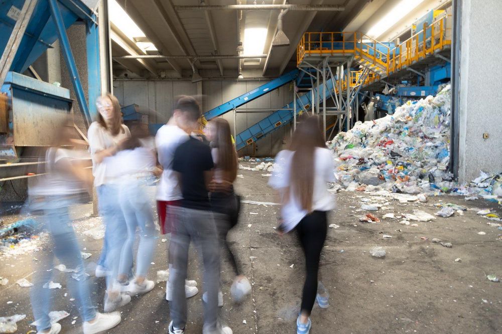 Un gruppo di persone balla in un impianto di riciclaggio, circondato da rifiuti e macchinari.