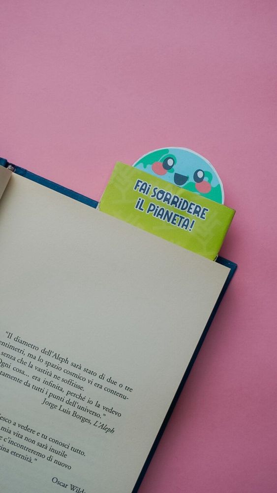 Un segnalibro con un messaggio che dice 'Fai sorridere il pianeta!' e un disegno di un pianeta con un volto felice, inserito in un libro aperto su uno