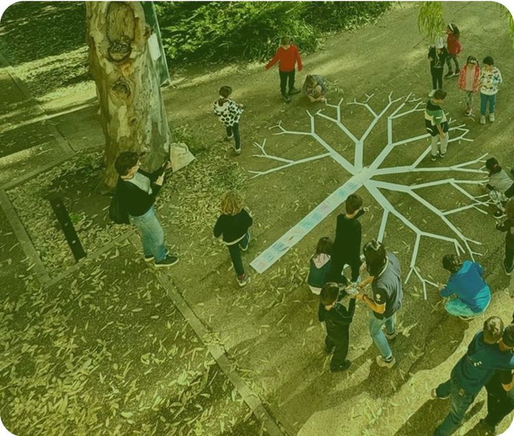 Un gruppo di persone, adulti e bambini, stanno intorno a un grande disegno di un albero fatto con del nastro adesivo colorato, su un terreno coperto d