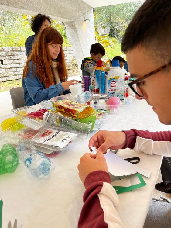 Bambini seduti intorno a un tavolo coperto di materiali riciclabili e oggetti di plastica, impegnati in un'attività manuale.