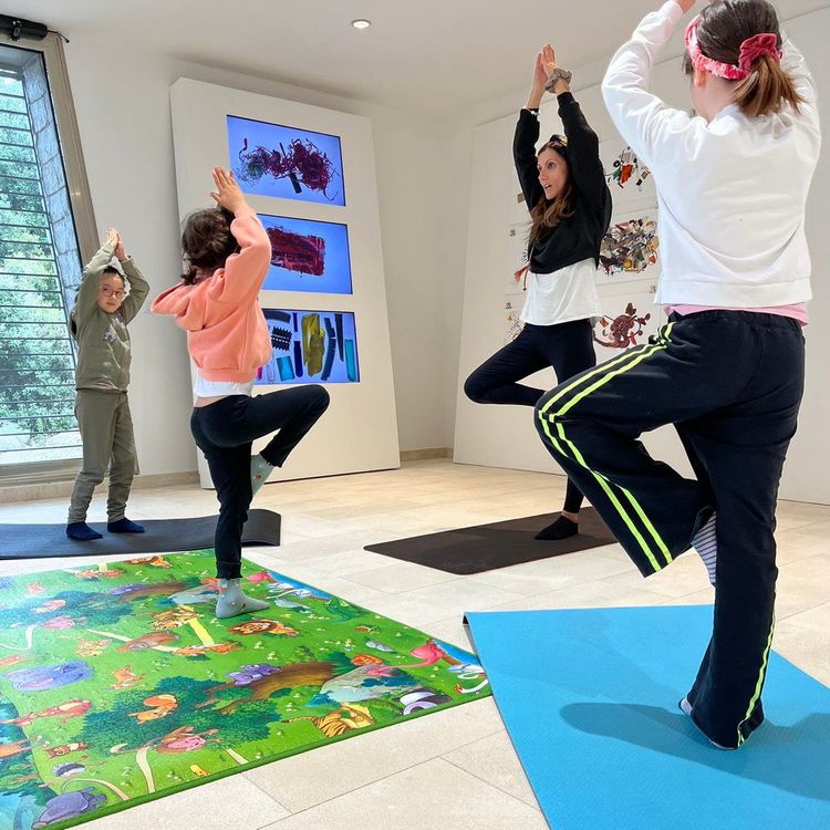 Quattro bambini, di varie età, praticano yoga su tappetini colorati in una stanza con pareti bianche e un pavimento in piastrelle beige, con una fines