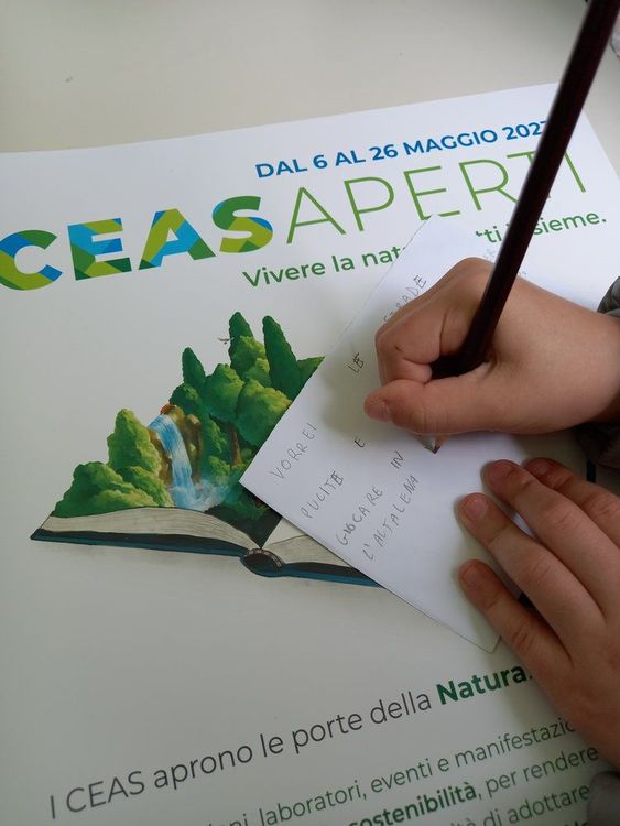 Mano di un bambino che scrive su un foglio con una matita, poggiato su un manifesto con un libro aperto e un paesaggio naturale