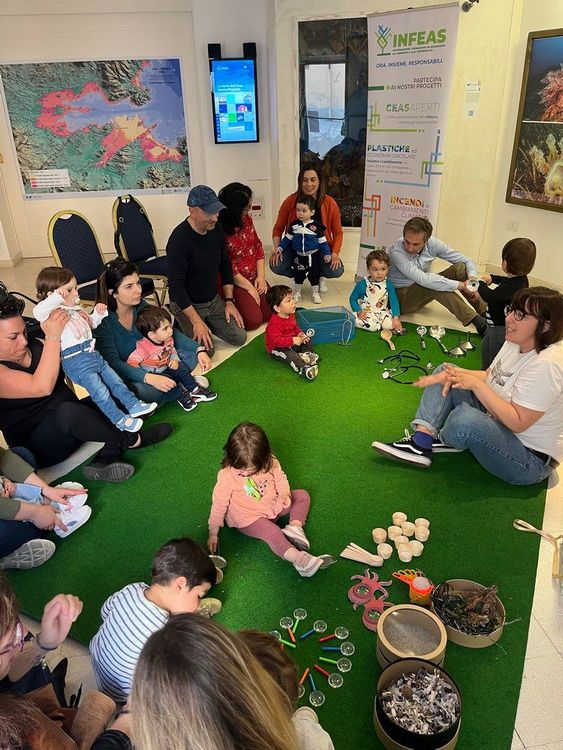 Un gruppo di adulti e bambini seduti in cerchio su un tappeto verde, con giocattoli e oggetti vari sparsi intorno a loro. Sembra un'attività educativa