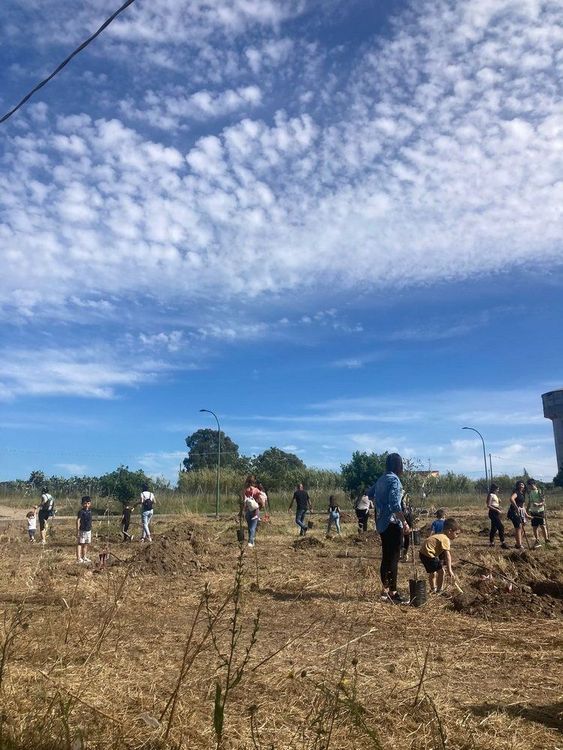 Gruppo di persone di varie età che lavorano insieme in un campo arido, sotto un cielo azzurro con nuvole bianche.