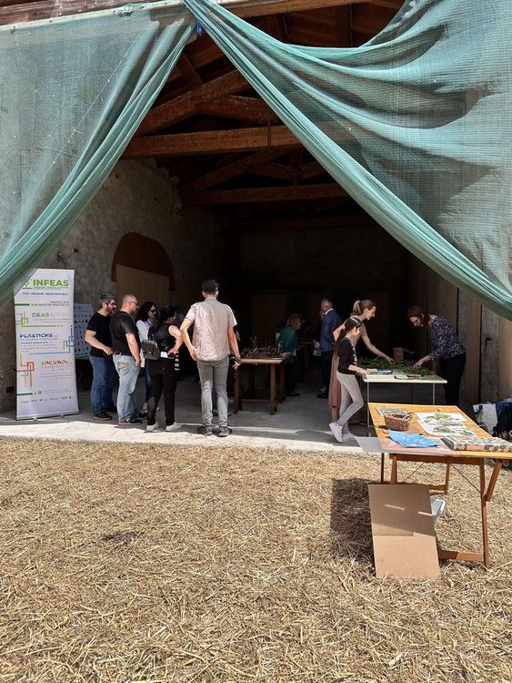 Un gruppo di persone entra in un capannone coperto da un telo, con un cartellone pubblicitario e un tavolino con materiali sul davanti