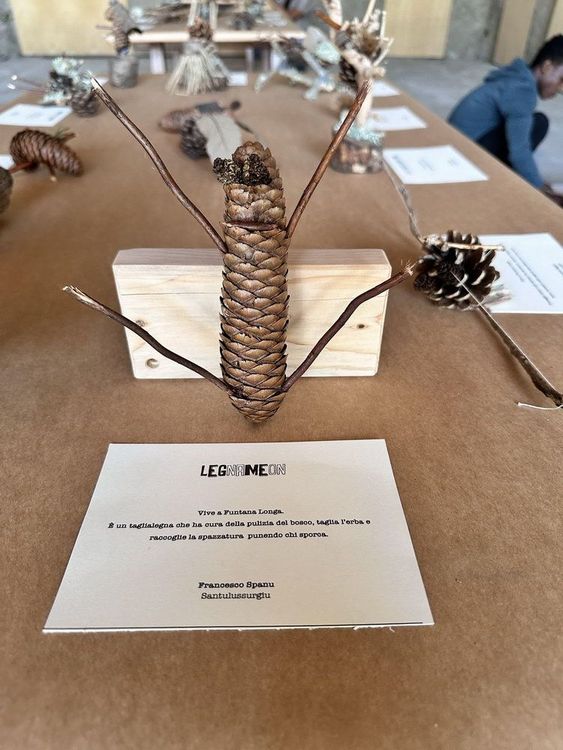 Scultura in legno e pigna su base di legno con cartellino descrittivo, su un tavolo con altre sculture simili.