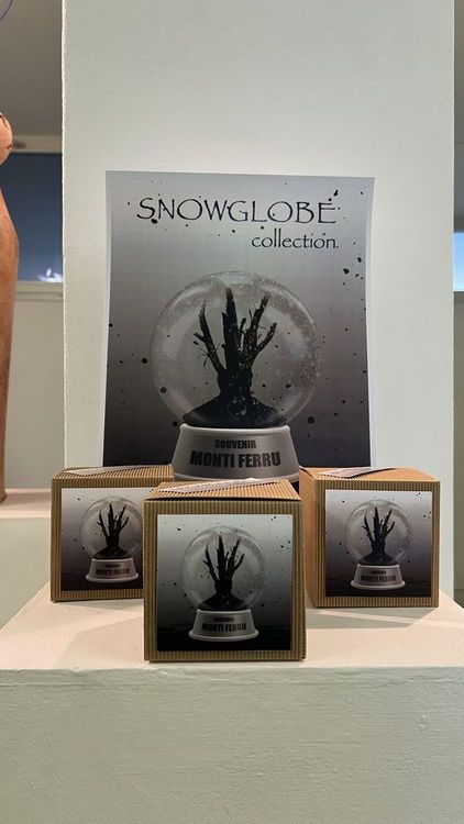 Tre scatole di souvenir della collezione Snowglobe con un albero nero all'interno di una palla di vetro, esposte su un ripiano.