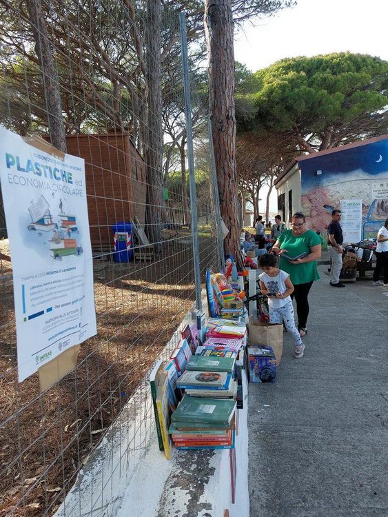 Una donna e un bambino passeggiano accanto a una recinzione con libri e un poster che promuove l'economia circolare e la riduzione delle plastiche.